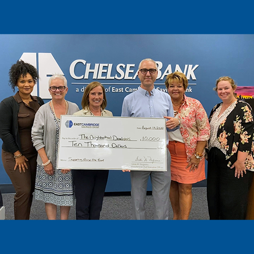 A group of representatives from the Bank, The Neighborhood Developers, and the Chelsea Chamber of Commerce stand for a photo with a ceremonial check. The photo was taken in front of a blue wall with the Bank's logo.